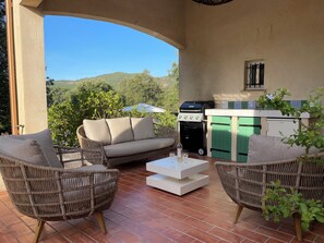 Eine Terrasse im Freien mit Korbmöbeln, einem Grill und Blick auf Grünflächen.