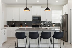 A modern kitchen with white cabinets, a central island with bar stools, a microwave, and a refrigerator. - 5BR Family Home, Pool, Theme Beds, Space, Location (Kissimmee)