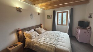 A bedroom with a wooden ceiling, a bed with a white blanket, a nightstand, a dresser, and a window with curtains. - Cosy village house (Jausiers)