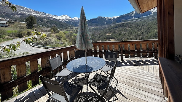 A wooden deck with a round table and chairs, overlooking a mountainous landscape. - Cosy village house (Jausiers)