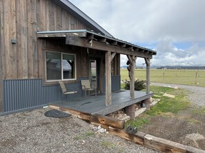 A rustic wooden building with a covered porch, two chairs, and a view of a grassy field and mountains.