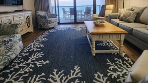 Ein Wohnzimmer mit einer Couch, einem Sessel, einem Couchtisch und einem Fernseher. Es gibt einen Balkon mit Stühlen und Blick zum Himmel.