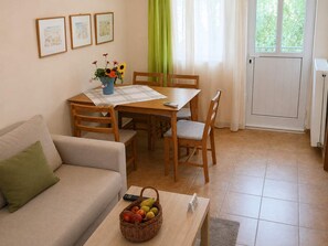 Een gezellige woonkamer met een bank, een eettafel met stoelen, een bijzettafeltje met fruit en een raam met gordijnen.