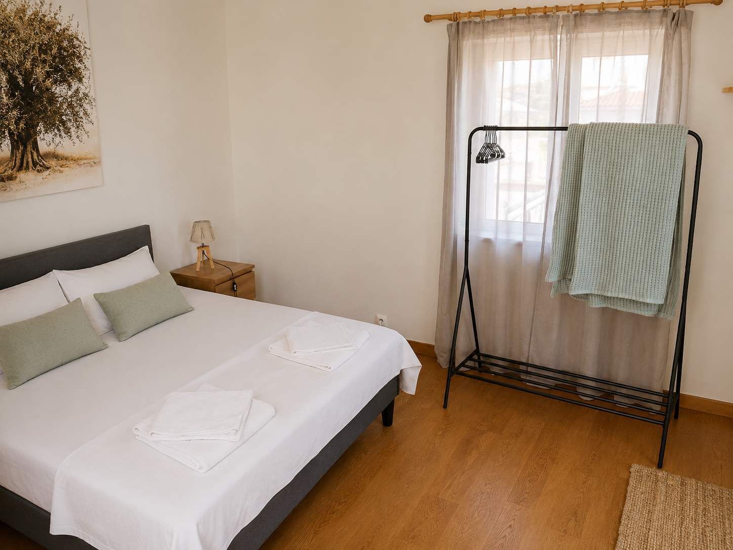 A neatly made bed with white linens, a bedside table with a lamp, and a window with sheer curtains.