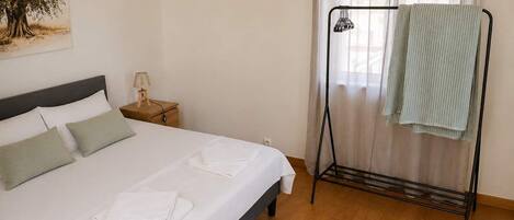 A neatly made bed with white linens, a bedside table with a lamp, and a window with sheer curtains.