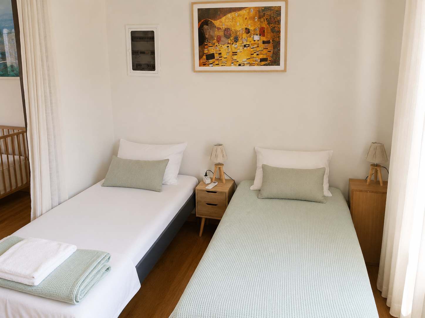 Une chambre d’hôtel avec deux lits simples, une table de chevet avec une lampe, un tableau au mur et une fenêtre avec des rideaux.