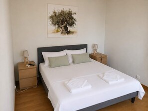Un llit ben fet amb roba blanca i dos coixins, una taula de nit amb una làmpada i un quadre penjat al mur que representa un arbre.