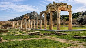 Ruinas antiguas con columnas de piedra y un arco parcialmente conservado, rodeado de campos verdes y un cielo despejado.