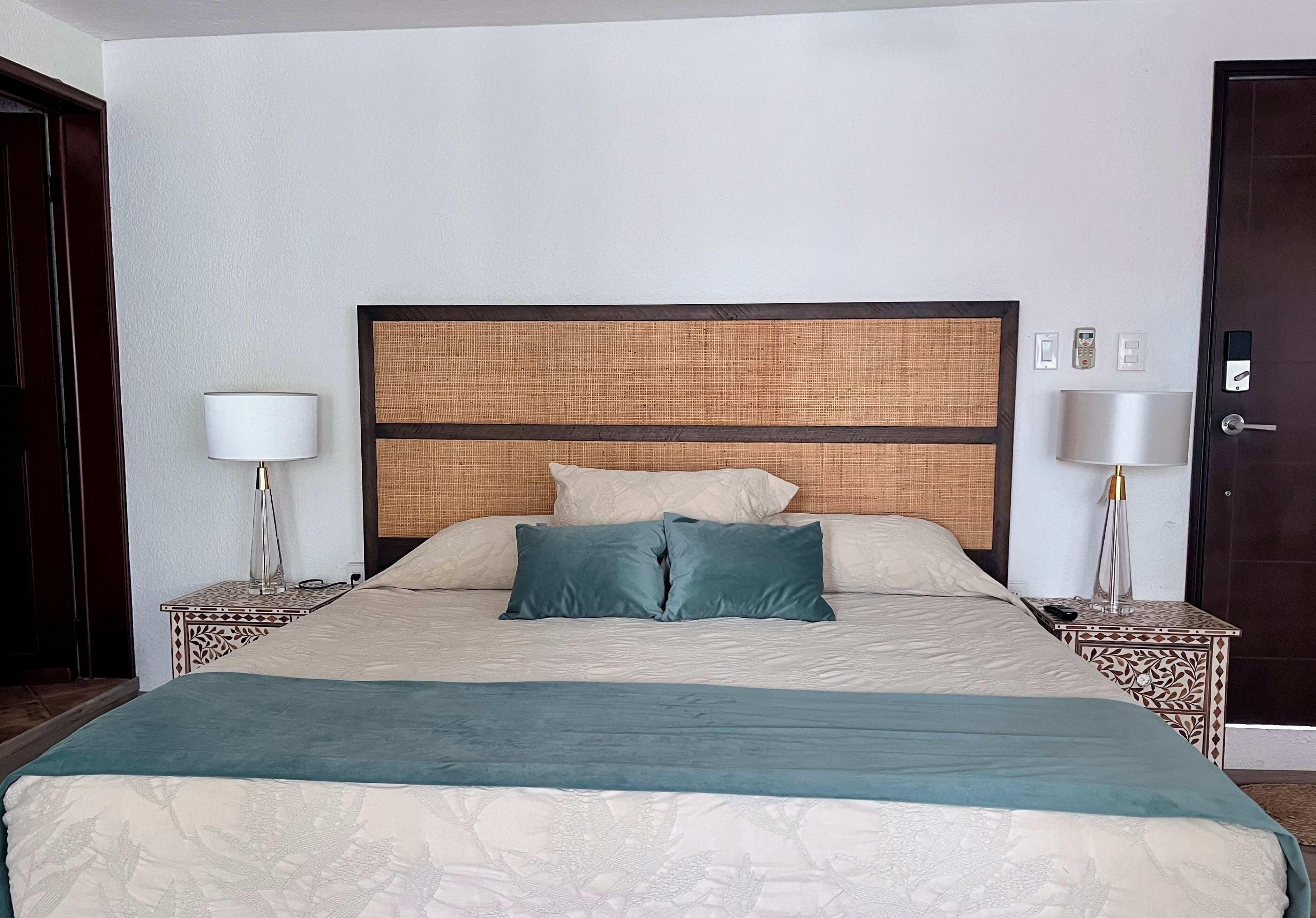 Un lit bien fait, recouvert d’une couvre-lit à motifs et agrémenté d’oreillers, flanqué de deux tables de chevet avec des lampes, dans une pièce dotée d’une porte en bois foncé et d’un mur sobre.