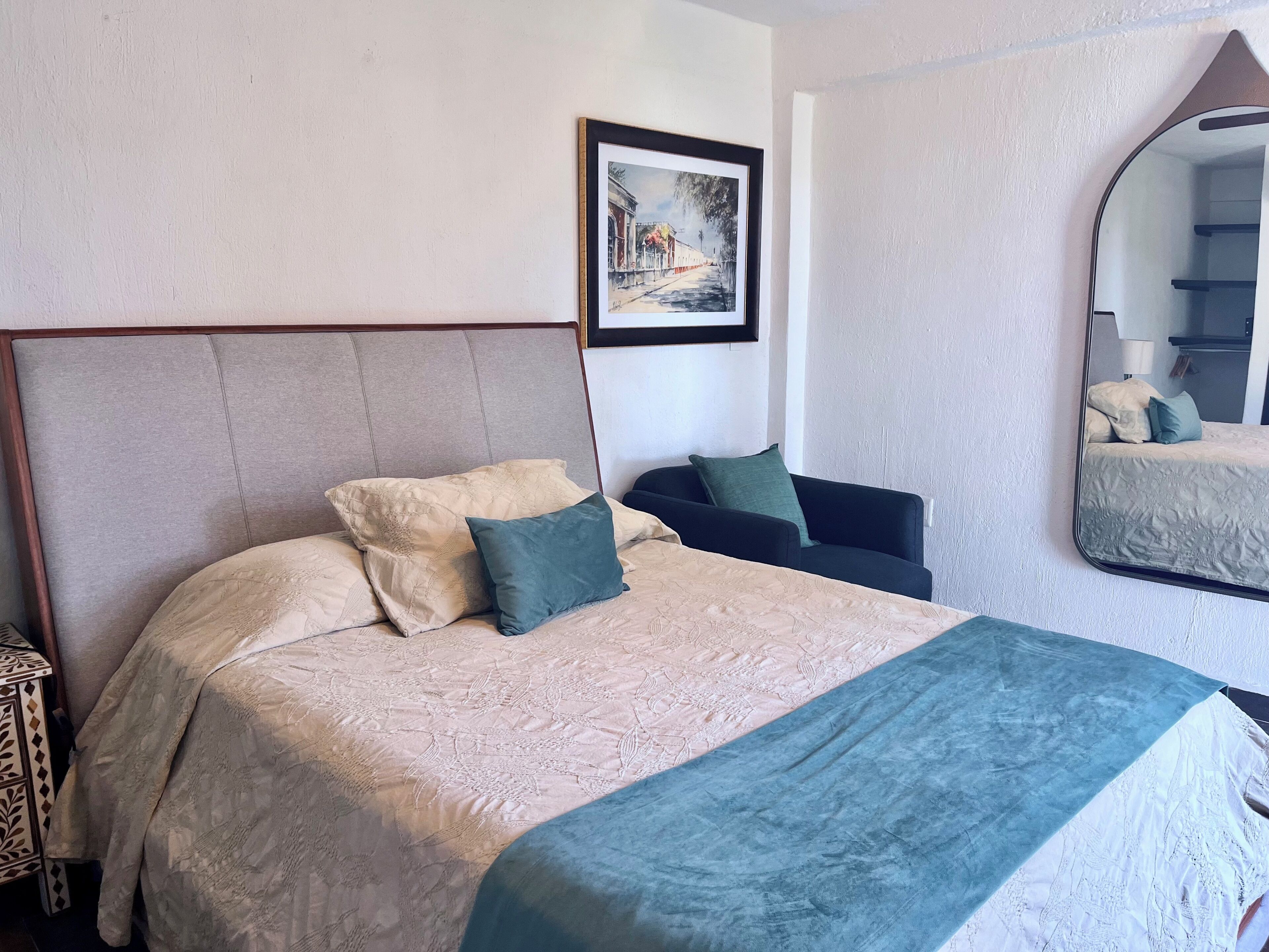 Una cama bien tendida con cabecera, una mesita de noche, una silla, un cuadro enmarcado en la pared y un espejo.