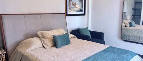 Una cama bien tendida con cabecera, una mesita de noche, una silla, un cuadro enmarcado en la pared y un espejo.