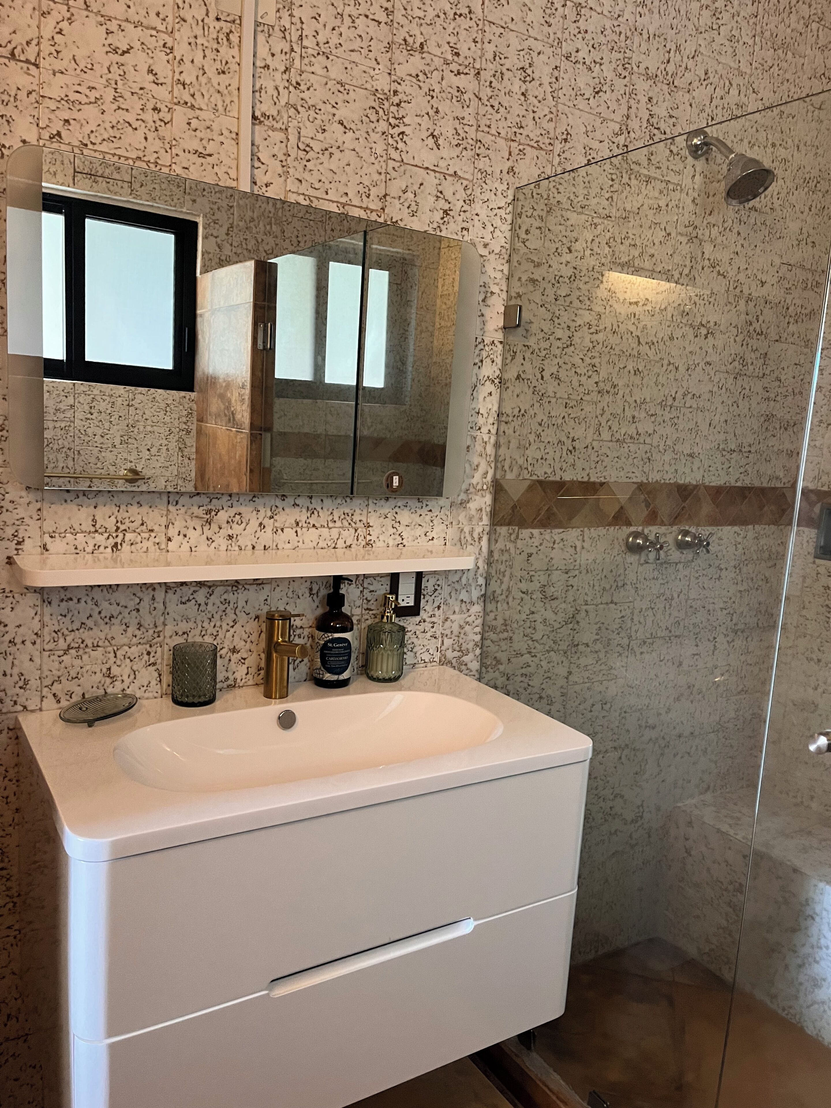 A bathroom with a white sink, a mirror above it, a glass shower enclosure, and a marble-patterned wall.