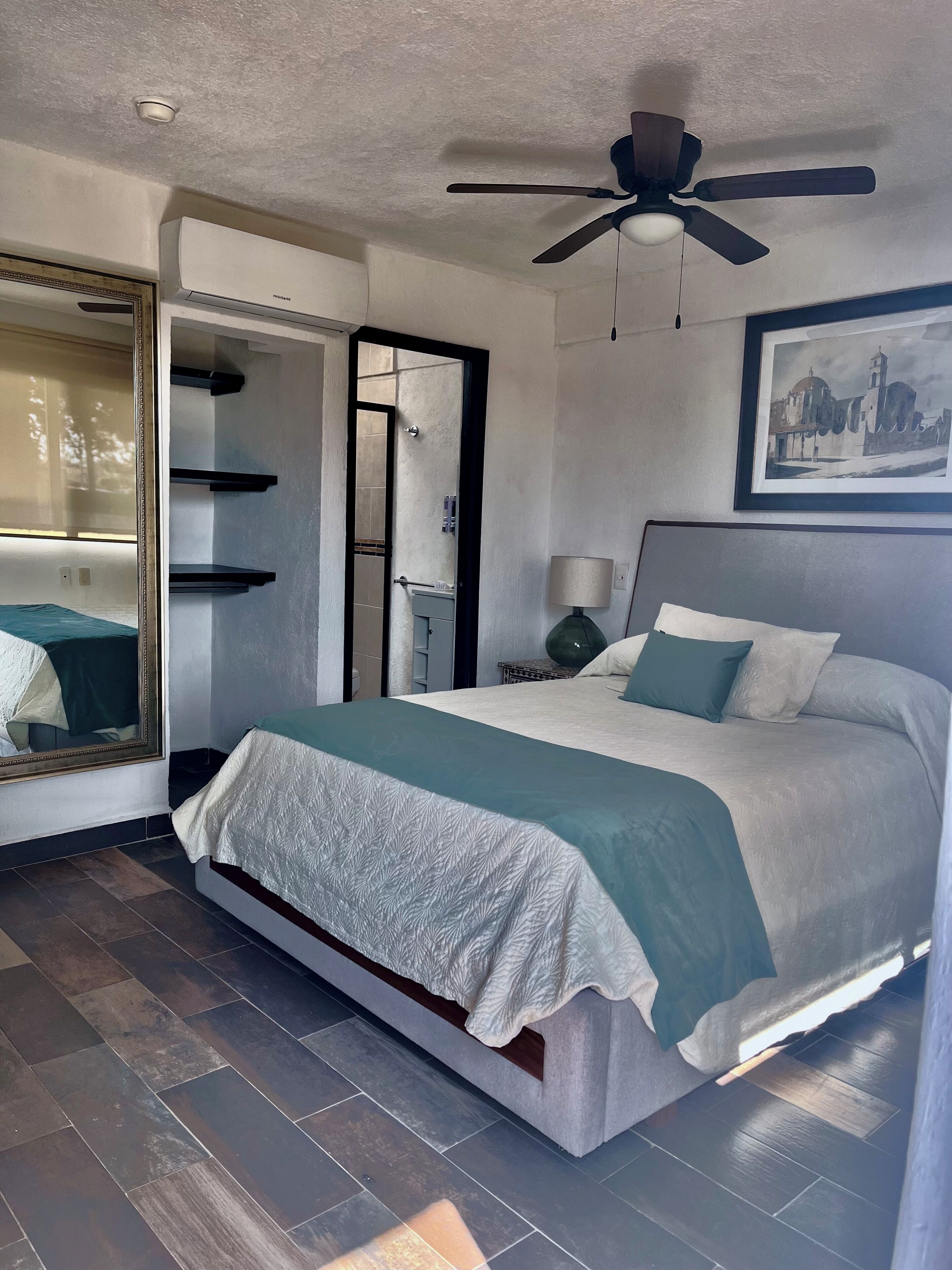 Un dormitorio con cama, ventilador de techo, un cuadro enmarcado y la puerta de un baño.
