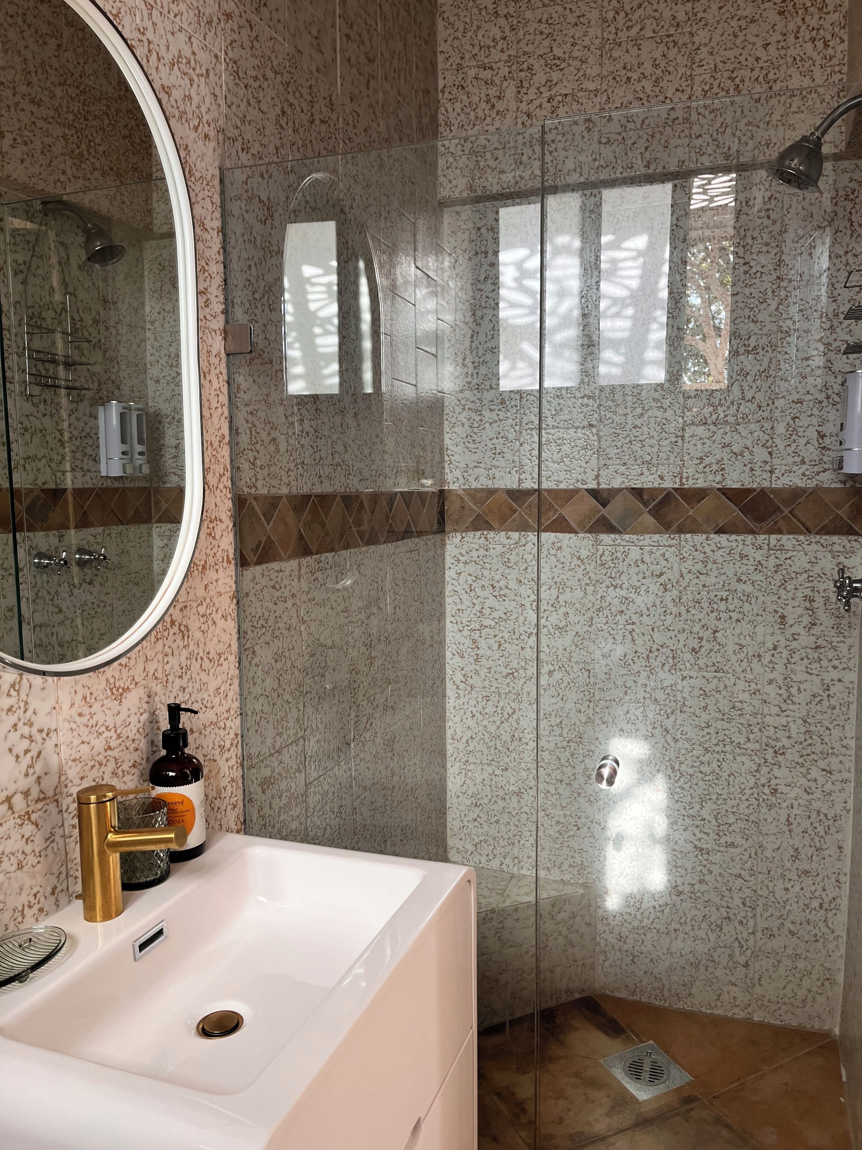 Une salle de bain avec un lavabo blanc, un miroir, une douche avec une porte en verre et un mur à motifs.