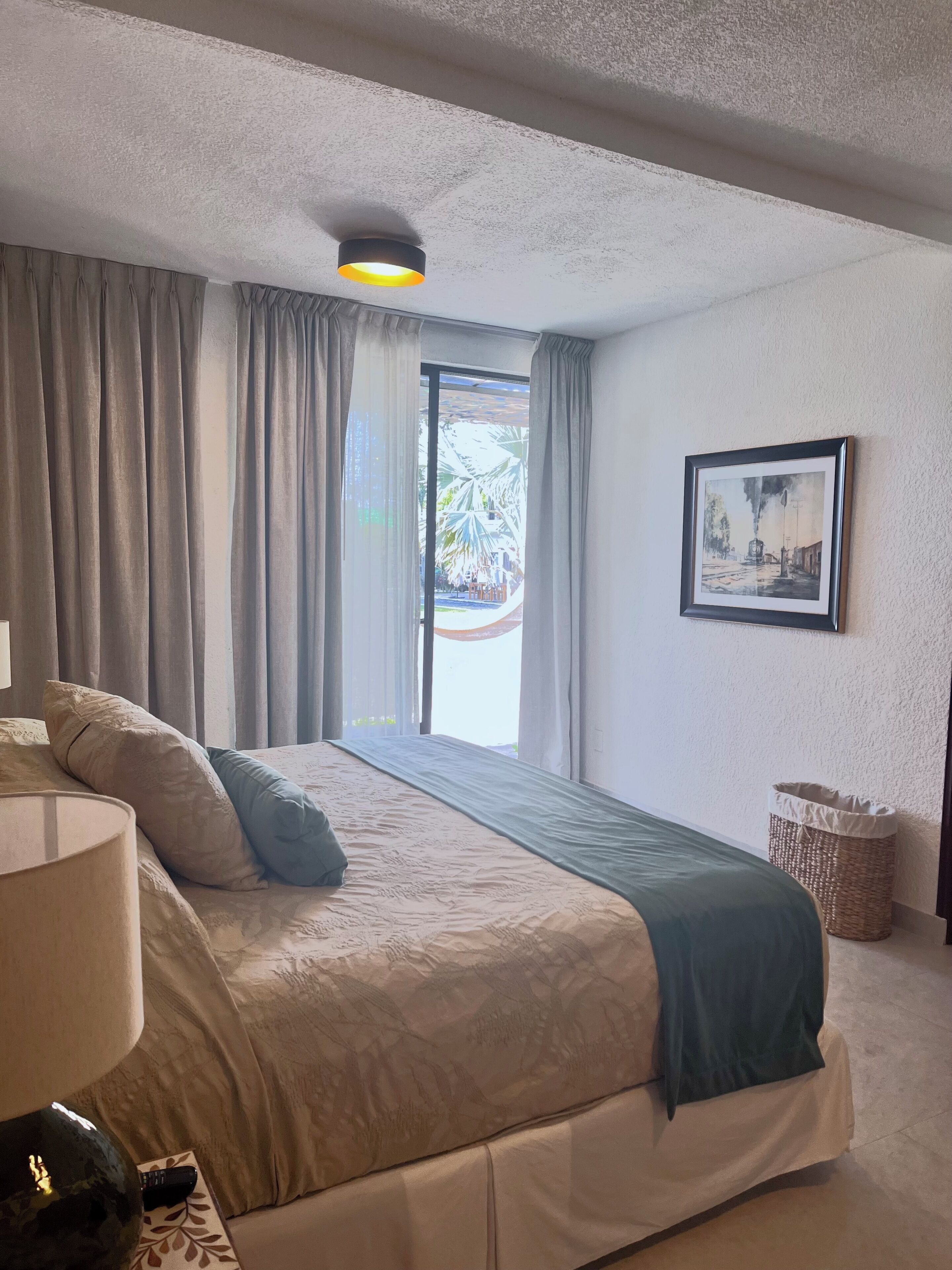 Un dormitorio con cama, cortinas, lámpara, un cuadro enmarcado y una cesta.