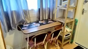 A room with a wooden desk, two chairs, a small shelf with a computer, and a white door. - Small Cosy Studio near City, Aldgate East (London)