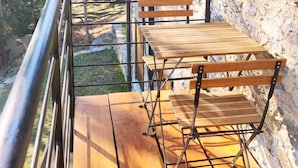 Een houten terras met een metalen hekwerk, een stenen gebouw met houten luiken en uitzicht op bomen.