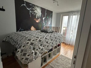 Een slaapkamer met een bedovertrek met dessin, een boekenkast, een groot muurschilderij en uitzicht op het landschap door een raam.