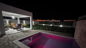 A swimming pool with stone flooring, a covered seating area with chairs, and a view of a residential area at night. - VILLA GREEN PARADISE GATED COMMUNITY SECURITY 24 HOURS  (puerto plata)