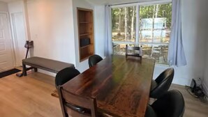 Tilava huone, jossa on suuri puinen pöytä, mustat tuolit, penkki ja suuret ikkunat, joista avautuu näkymä metsään.