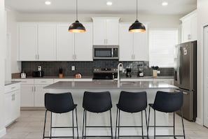 A modern kitchen with white cabinets, a central island with bar stools, a microwave, and a refrigerator. - 5BR Family Home, Pool, Theme Beds, Space, Location (Kissimmee)