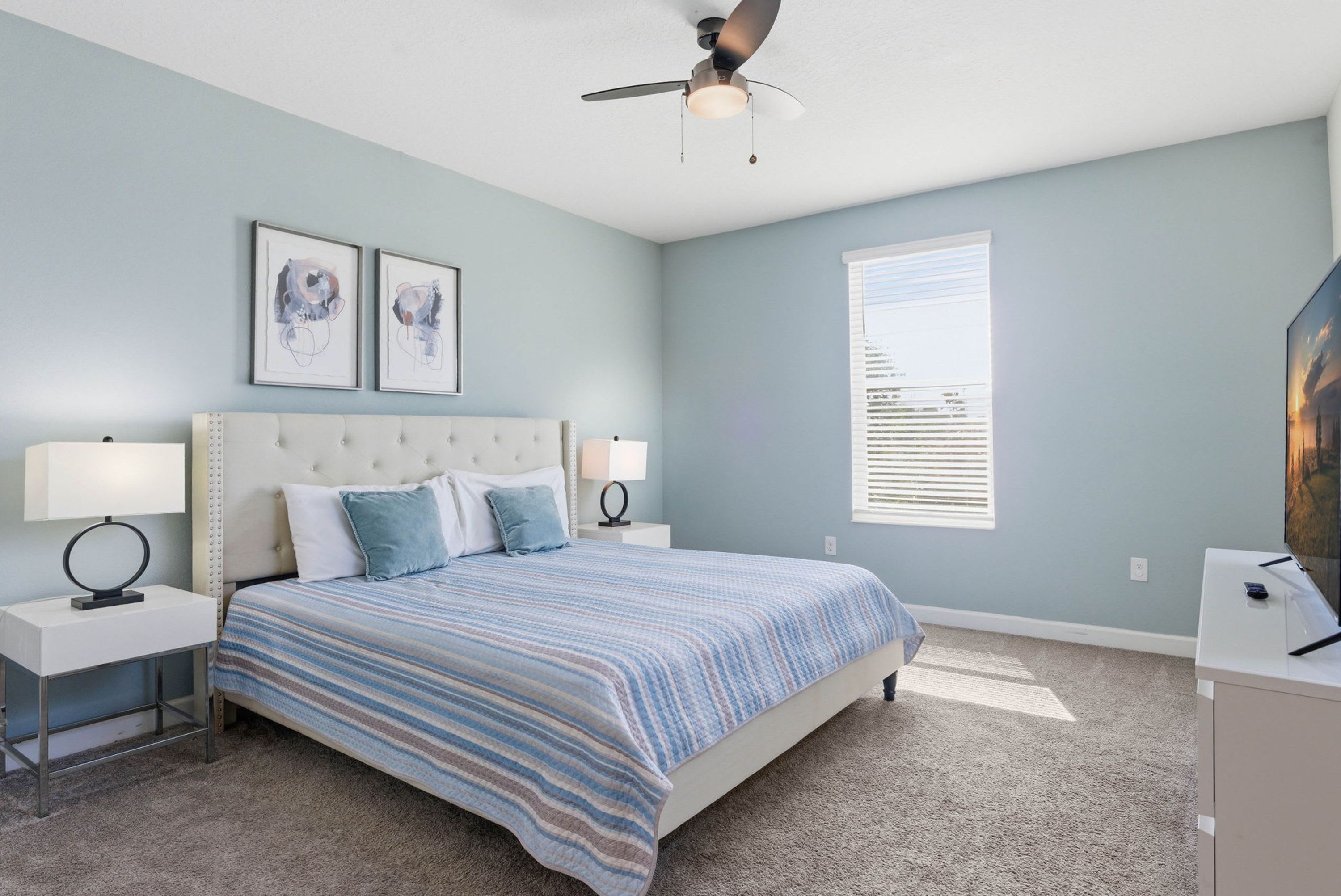 A bedroom with a bed, nightstands, a ceiling fan, a window, and artwork on the wall.