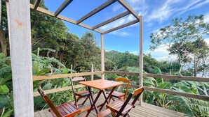 A wooden deck with a table and chairs, surrounded by lush greenery and a clear sky. - At the top of Tainha beach, with a beautiful view that you can only find at Cabana do Abib. (Bombinhas)