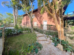 A garden with a stone pathway, a wooden deck, and a house with a red exterior. - At the top of Tainha beach, with a beautiful view that you can only find at Cabana do Abib. (Bombinhas)