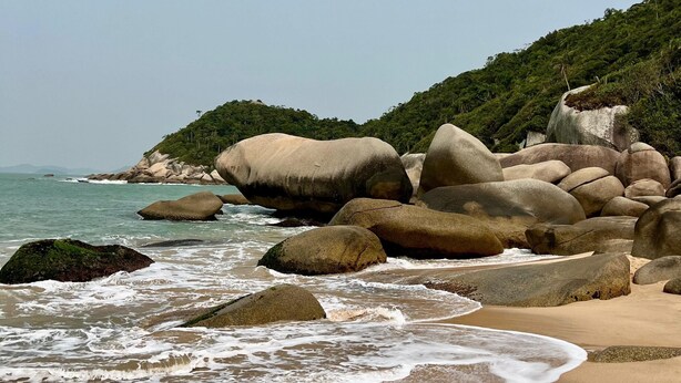 A beach with large boulders, calm waves, and a clear sky. - At the top of Tainha beach, with a beautiful view that you can only find at Cabana do Abib. (Bombinhas)