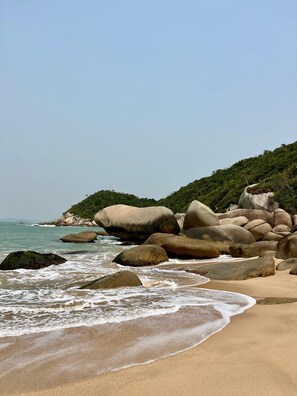 Ein Strand mit großen Felsen, ruhiger Brandung und klarem Himmel.