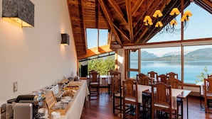 Un comedor con mesas y sillas de madera, un bufé con comida y bebidas, y vistas a un lago y montañas a través de amplias ventanas.