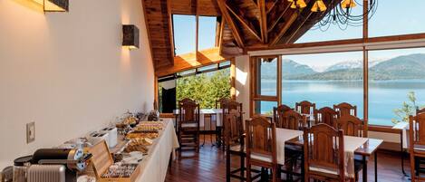 A dining area with wooden tables and chairs, a buffet with food and drinks, and a view of a lake and mountains through large windows.