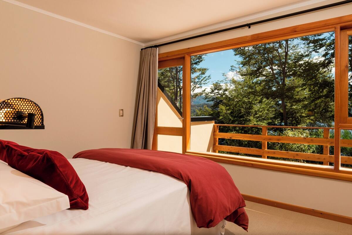 Quarto com uma janela grande, uma cama com roupa de cama vermelha e branca, e uma cabeceira de madeira.