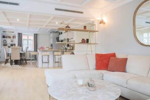 Ein modernes Wohnzimmer mit einem weißen Sofa, einem Couchtisch sowie einem Essbereich mit Stühlen und einer Küche im Hintergrund.