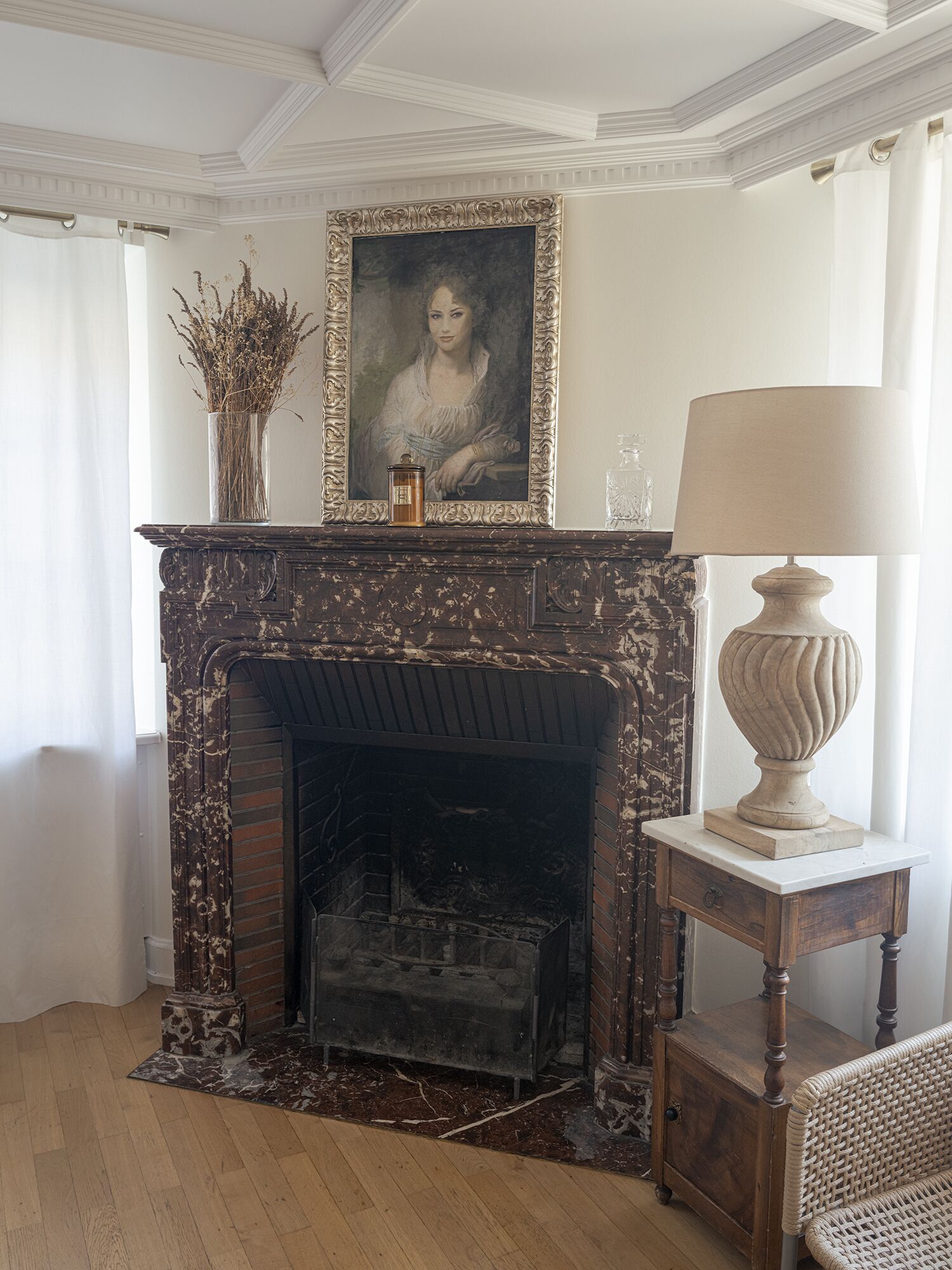 Une pièce avec une cheminée, un portrait encadré, un vase contenant des plantes séchées, une lampe et un fauteuil en osier.