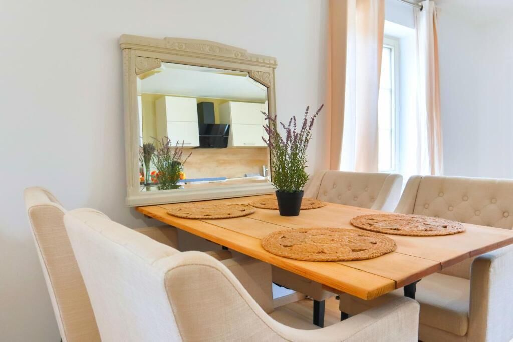 A dining area with a wooden table, beige upholstered chairs, a large mirror, and a potted plant.