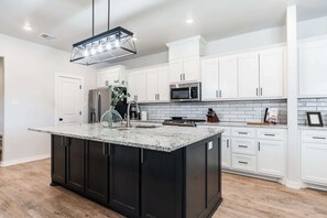 A modern kitchen with white cabinets, a black island, stainless steel appliances, and a geometric pendant light. - The Carlisle - 3BR/2BA - Family Friendly New Build (Lubbock)