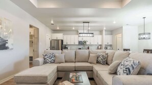 A modern living room with a sectional sofa, a coffee table, and a kitchen in the background. - The Carlisle - 3BR/2BA - Family Friendly New Build (Lubbock)