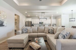 A modern living room with a sectional sofa, a coffee table, and a kitchen in the background. - The Carlisle - 3BR/2BA - Family Friendly New Build (Lubbock)