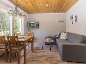 A cozy room with a wooden ceiling, a dining table with chairs, a gray sofa, a small coffee table, and a TV mounted on the wall.