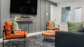Una sala de estar moderna con un televisor de pantalla plana montado en la pared, dos sillones de cuero con cojines, una mesa de centro de madera y una alfombra estampada.