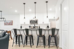A modern kitchen with white cabinets, a stainless steel microwave, and a bar area with four stools. - Hidden Lake Gem Arcade Stay (Denison)