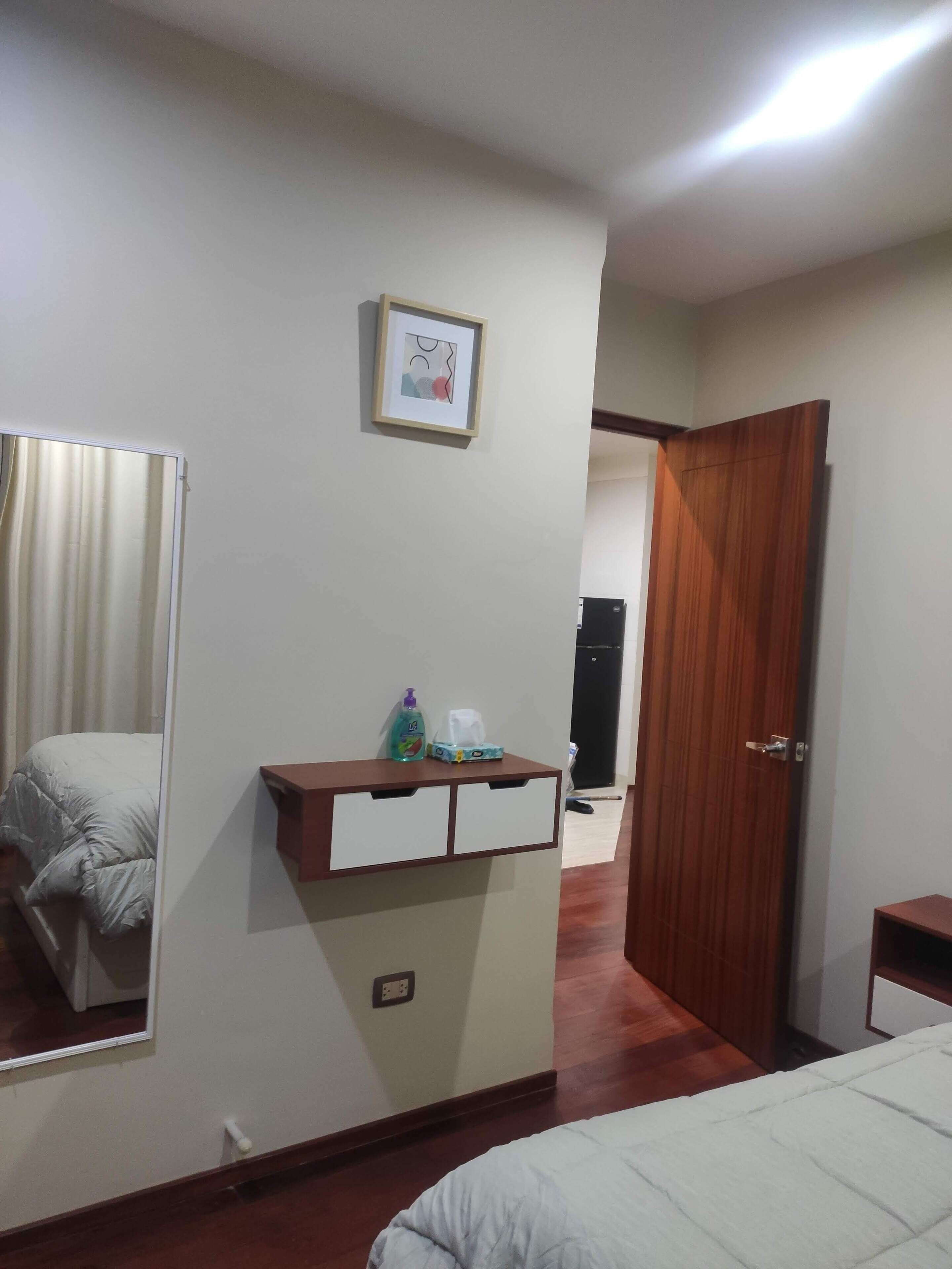 A bedroom with a bed, a bedside table with a mirror, a framed picture on the wall, and a door leading to another room.