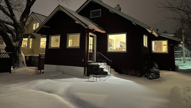 Una casa de madera oscura con varias ventanas, una escalera visible y el suelo cubierto de nieve.