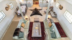 Une pièce spacieuse avec un grand tapis en peau d’ours, deux canapés verts ornés de coussins à motifs, une table basse en bois et un coin repas avec une nappe blanche.