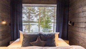 A bed with white bedding and two pillows, a window with a view of trees and a body of water, and wooden walls with wall sconces.