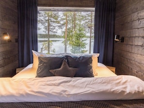 Una cama con ropa de cama blanca y dos almohadas, una ventana con vista a árboles y un cuerpo de agua, y paredes de madera con apliques.