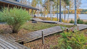 Une terrasse en bois mène à un bâtiment doté d’un espace extérieur couvert, le tout étant entouré d’arbres et bordé par un lac.