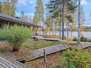 Una terraza de madera que conduce a un edificio con un espacio al aire libre cubierto, rodeado de árboles y un lago.