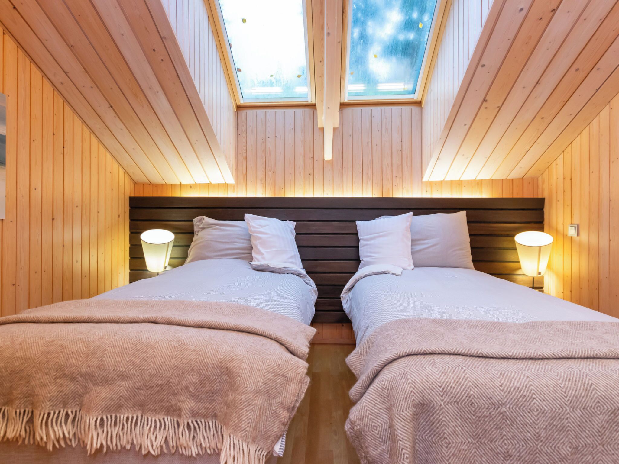 Une pièce aux murs en bois, un grand lit avec deux oreillers, une table de chevet avec une lampe et une lucarne au plafond.